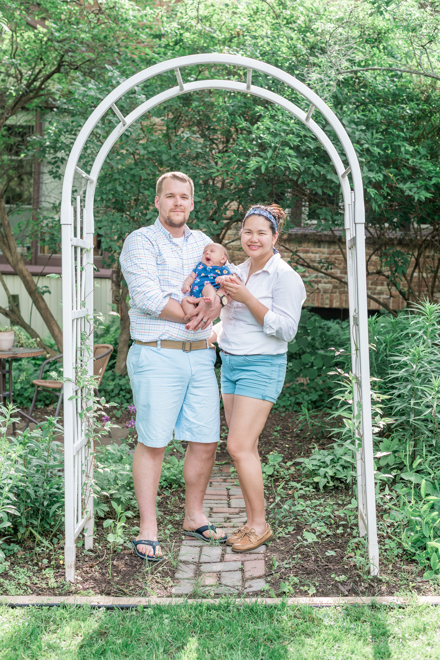 Father and mother smiles with newborn baby girl under their backyard arch