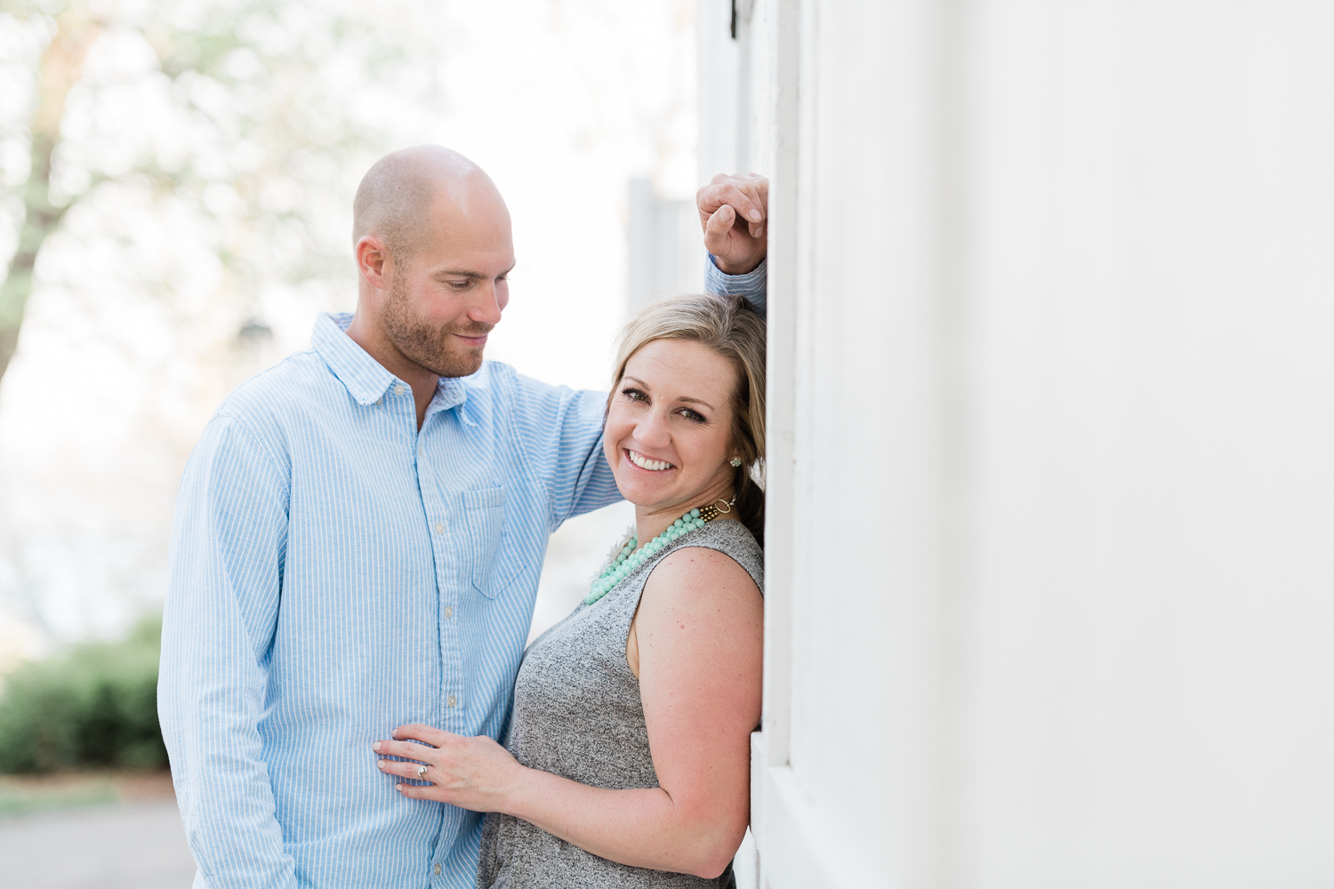 Fiance unbuttons light blue shirt in front of giggling fiance against a white wall