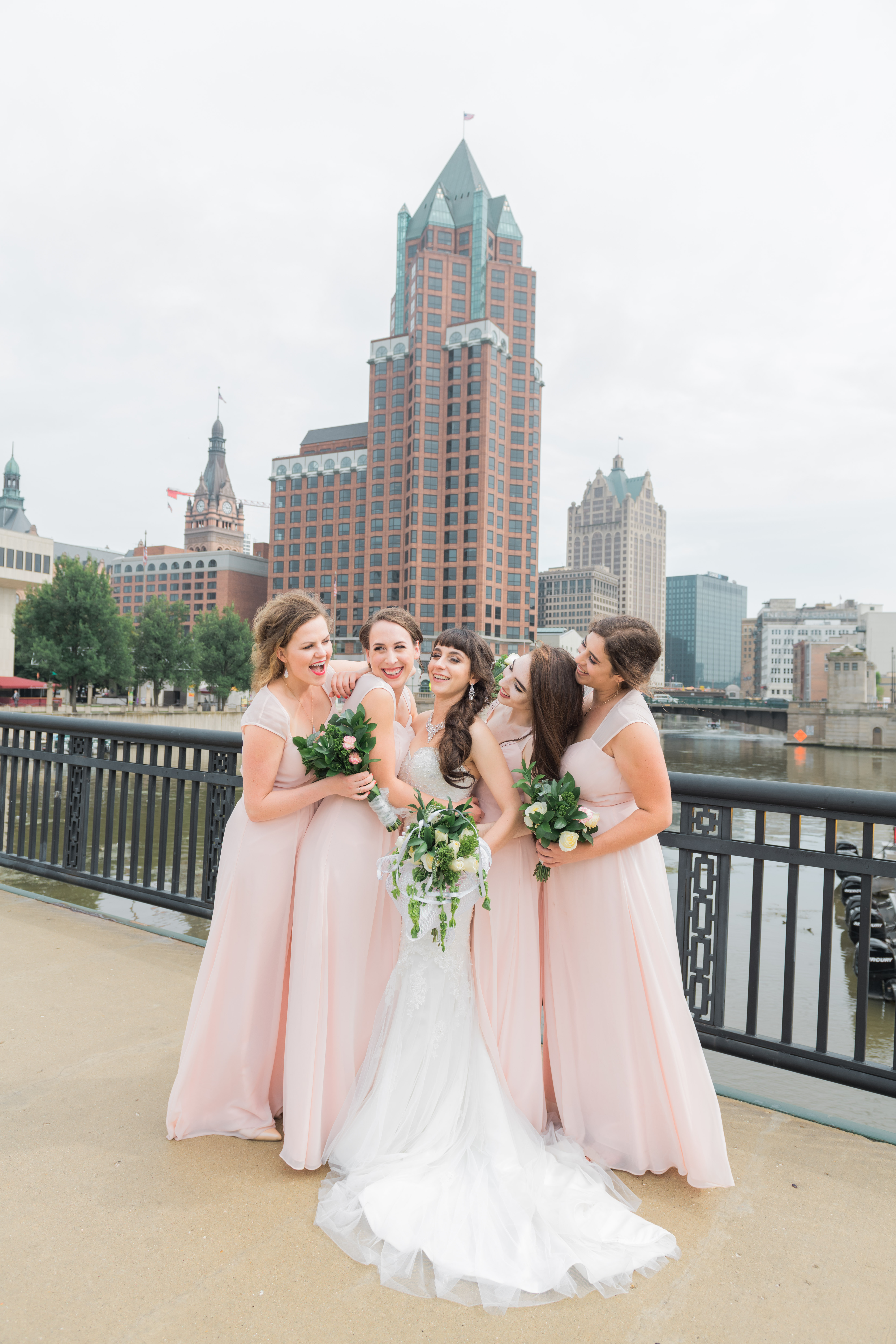 Bridesmaids surround bride smiling at each other by the Milwaukee bridge with large building in the background