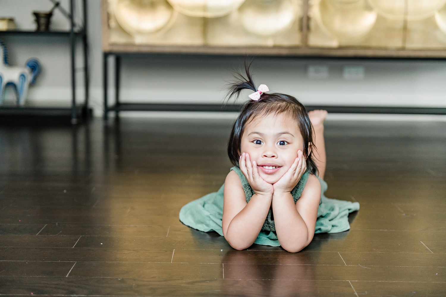 Toddler in green dress and small pink bow lies down on living room floor with hands catching her cheeks