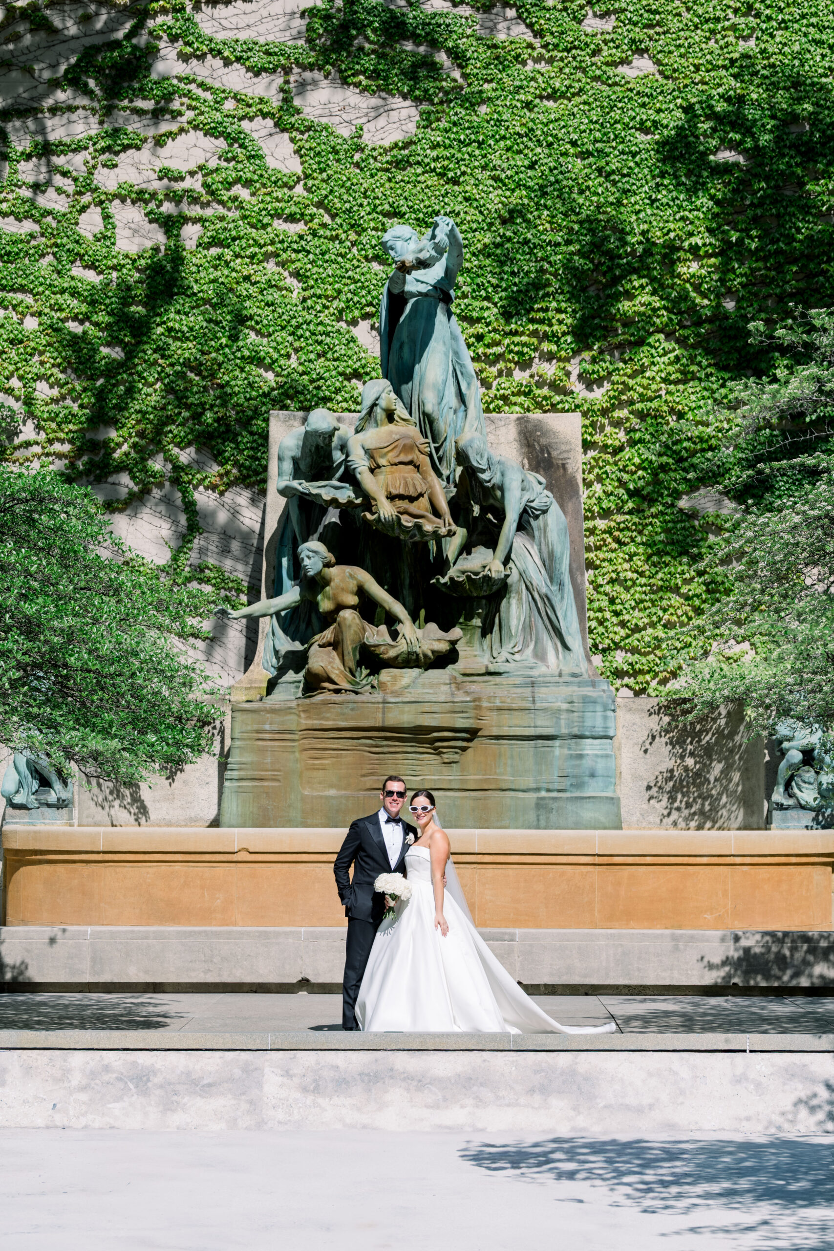 A bride and groom in formal wedding attire pose in front of a grand ivy-covered fountain with classical sculptures, captured in bright natural light.